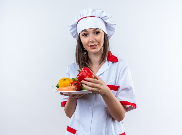 Pleased young female cook wearing chef uniform holding vegetables on plate isolated on white wall