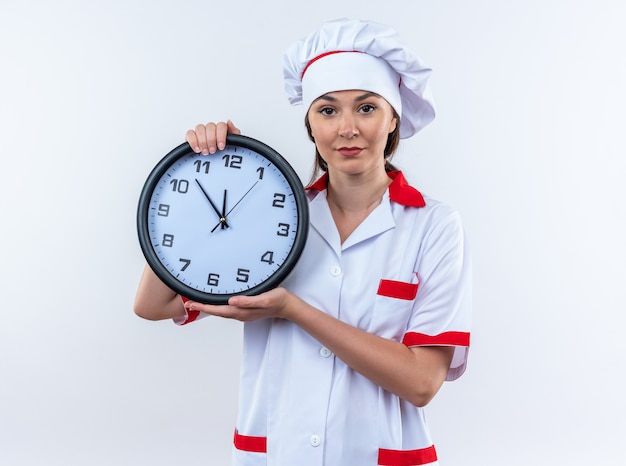 Foto gratuita piacere giovane cuoca che indossa l'uniforme dello chef tenendo l'orologio da parete isolato su parete bianca