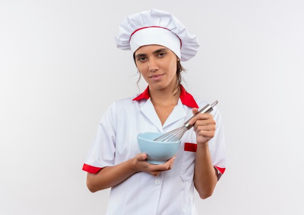 довольная молодая женщина-повар в униформе шеф-повара держит венчик и миску на изолированной белой стене с копией пространства