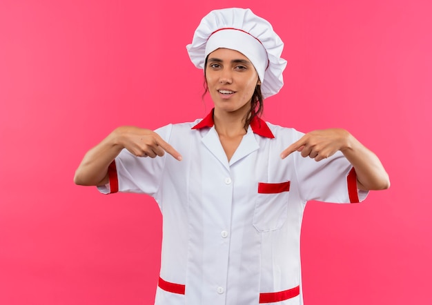 Pleased young female cook wearing chef uniform points to herself with copy space