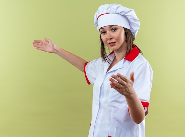 オリーブグリーンの背景で隔離の何かを保持しているふりをしてシェフの制服を着て喜んで若い女性料理人