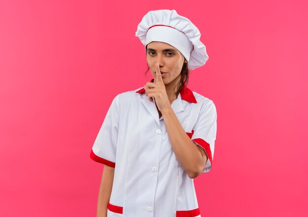  pleased young female cook wearing chef uniform showing silence gesture on isolated pink wall with copy space