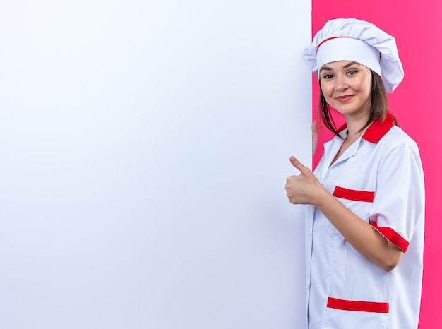 シェフの制服を着て満足している若い女性料理人は、コピースペースとピンクの背景で隔離の親指を示す白い壁の近くに立っています