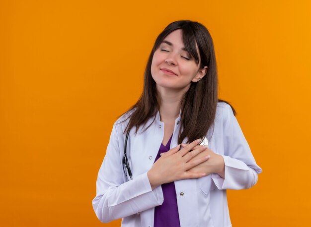 Pleased young female doctor in medical robe with stethoscope holds heart with both hands on isolated orange background with copy space