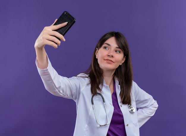 청진 기 의료 가운에 기쁘게 젊은 여성 의사는 전화를 보유하고 격리 된 보라색 배경에 전화를 찾습니다
