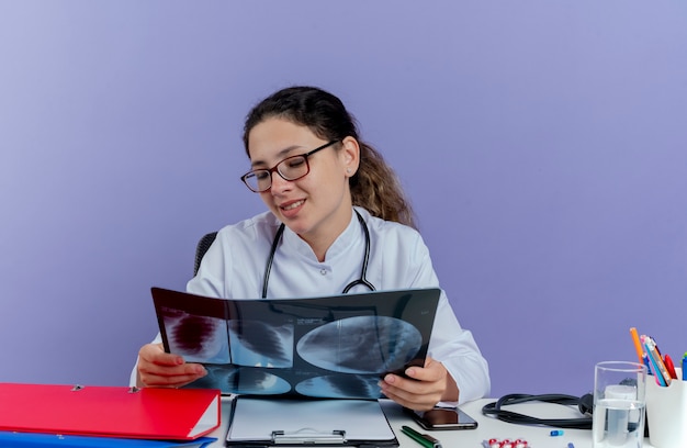 Бесплатное фото Довольная молодая женщина-врач в медицинском халате и стетоскопе сидит за столом с медицинскими инструментами и смотрит на изолированный рентгеновский снимок