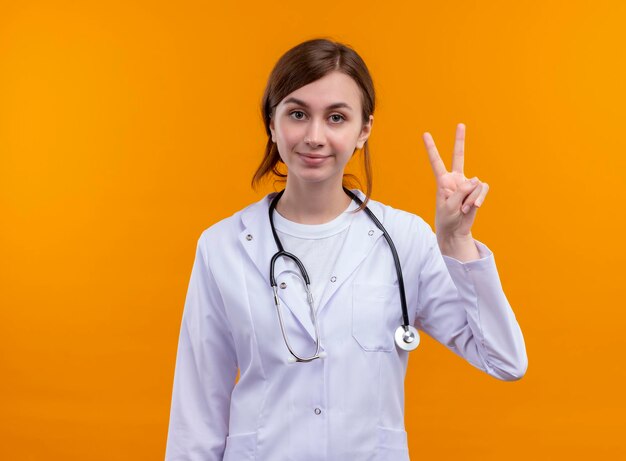 Pleased young female doctor wearing medical robe and stethoscope doing peace sign on isolated orange space with copy space