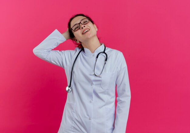 Pleased young female doctor wearing medical robe and stethoscope and glasses putting hand behind head with closed eyes isolated on pink wall with copy space