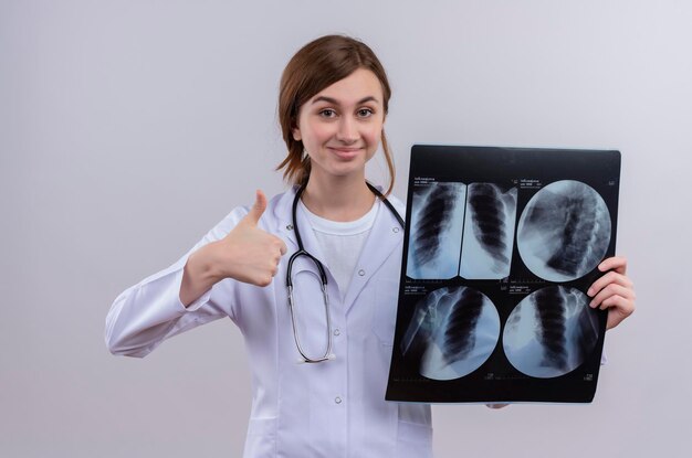 Pleased young female doctor wearing medical robe and stethoscope and holding x-ray shot showing thumb up 