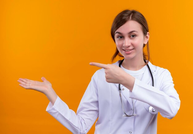 Pleased young female doctor wearing medical robe and stethoscope pointing at left side and showing empty hand on isolated orange space