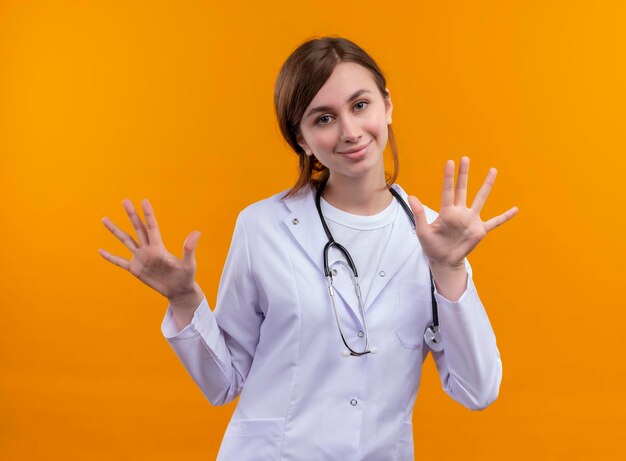 Pleased young female doctor wearing medical robe and stethoscope and showing empty hands on isolated orange space