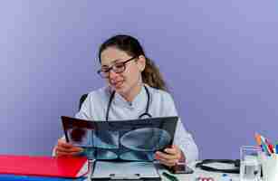 Free photo pleased young female doctor wearing medical robe and stethoscope sitting at desk with medical tools holding and looking at x-ray shot isolated