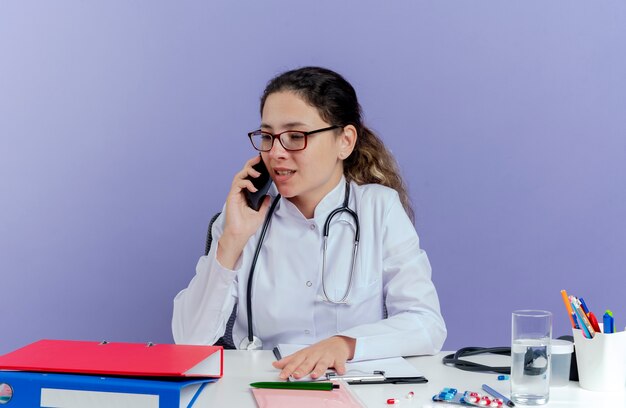 Pleased young female doctor wearing medical robe and stethoscope sitting at desk with medical tools looking at side talking on phone isolated