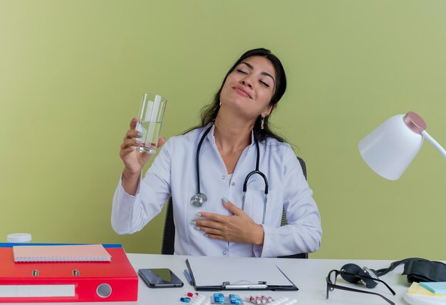 Довольная молодая женщина-врач в медицинском халате и стетоскопе сидит за столом с медицинскими инструментами, положив руку на грудь, держа стакан воды с закрытыми изолированными глазами