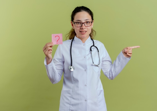 Pleased young female doctor wearing medical robe and stethoscope with glasses holding paper note points at side isolated on olive green wall with copy space