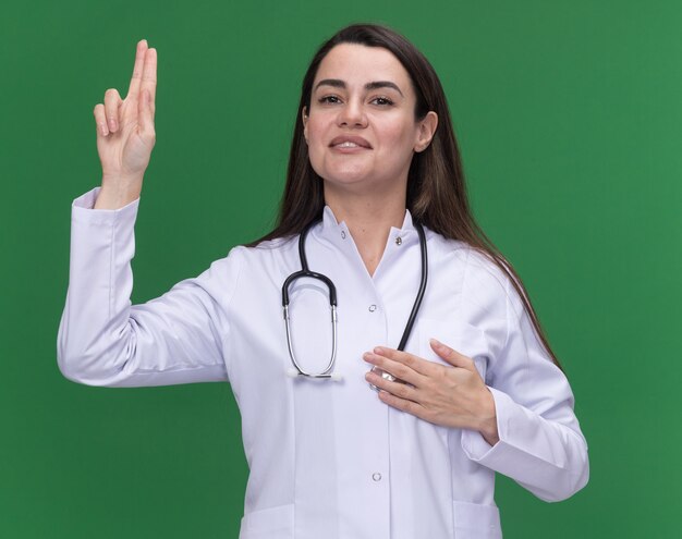 Довольная молодая женщина-врач в медицинском халате со стетоскопом делает жест клятвы на зеленом