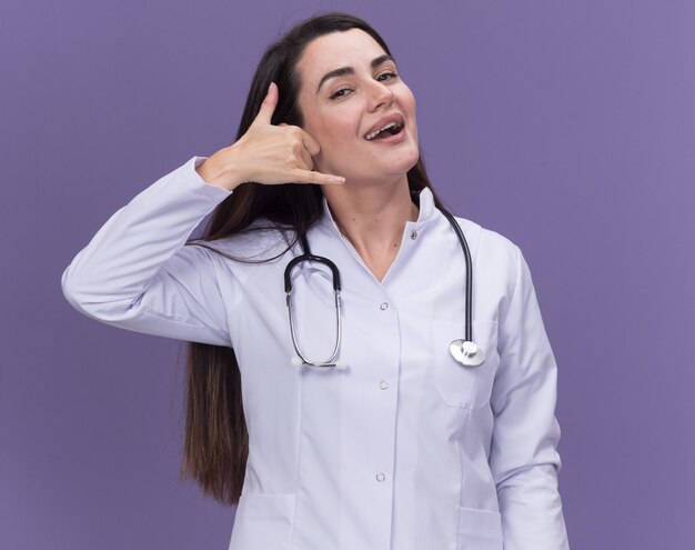 Довольная молодая женщина-врач в медицинском халате со стетоскопом жесты позвоните мне, изолированные на фиолетовой стене с копией пространства