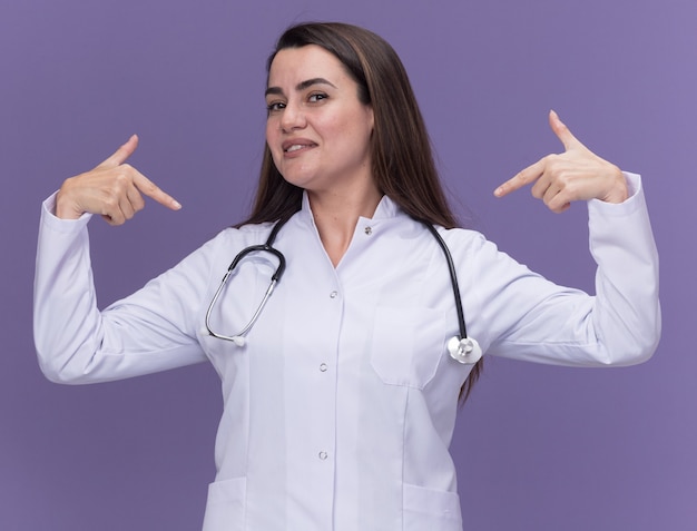 Pleased young female doctor wearing medical robe with stethoscope points at herself with two hands on purple 