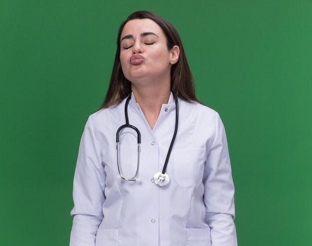 Pleased young female doctor wearing medical robe with stethoscope pretends to kiss on green 
