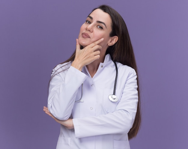 Lieta giovane dottoressa che indossa una veste medica con lo stetoscopio mette la mano sul mento e guarda la telecamera sul viola