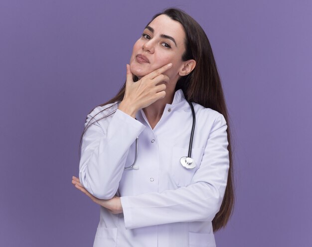 Pleased young female doctor wearing medical robe with stethoscope puts hand on chin and looks at camera on purple 