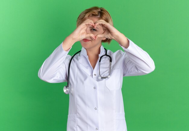 Pleased young female doctor wearing medical robe with stethoscope showing heart gesture isolated on green wall