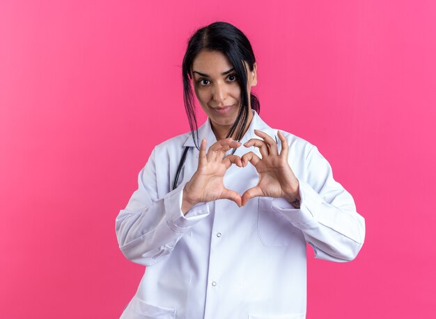 Довольная молодая женщина-врач в медицинском халате со стетоскопом показывает жест сердца, изолированный на розовой стене