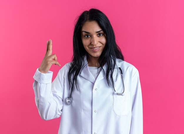 Довольная молодая женщина-врач в медицинском халате со стетоскопом показывает жест пистолета, изолированный на розовой стене