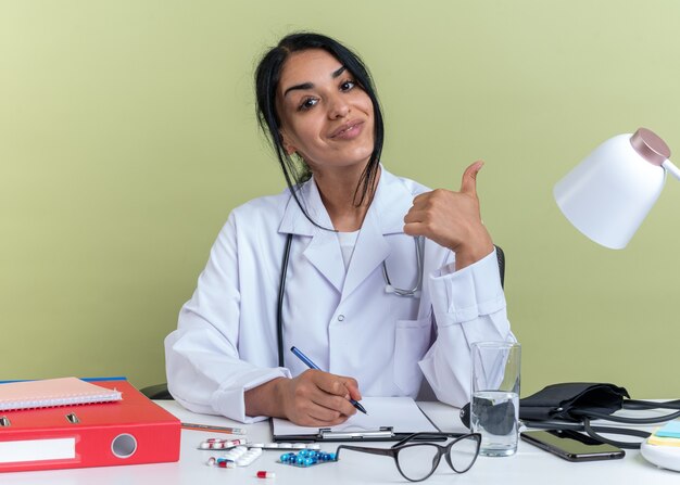 Pleased young female doctor wearing medical robe with stethoscope sits at desk with medical tools writing something on clipboard showing thumb up isolated on olive green wall