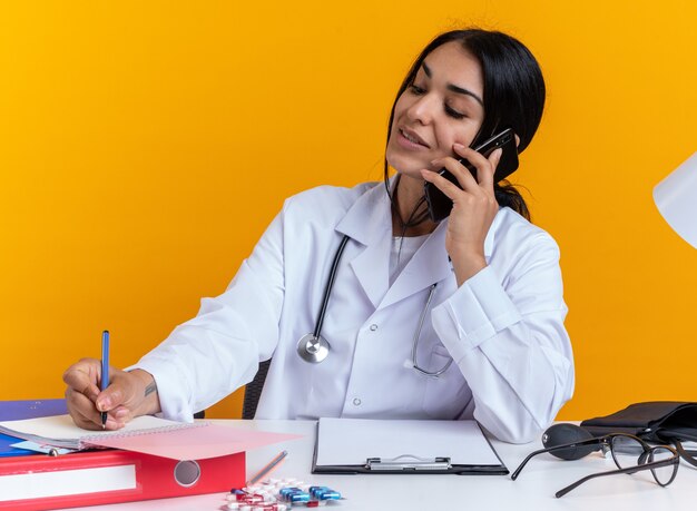 청진 기 의료 가운을 입고 기쁘게 젊은 여성 의사 의료 도구 테이블에 앉아 노란색 벽에 고립 된 노트북에 뭔가 쓰는 전화에 말한다