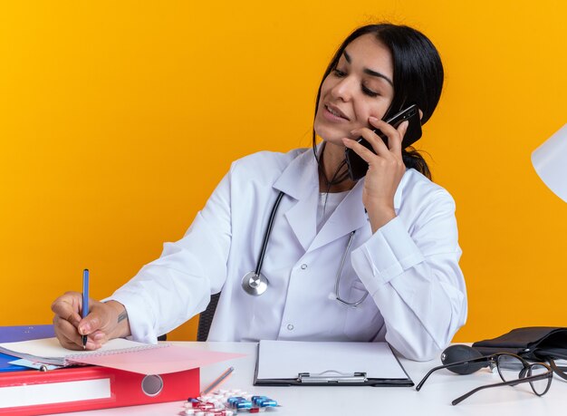 Pleased young female doctor wearing medical robe with stethoscope sits at table with medical tools speaks on phone writing something on notebook isolated on yellow wall