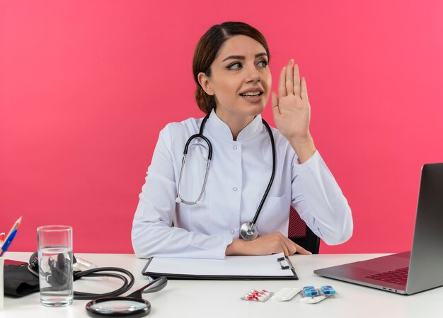 Pleased young female doctor wearing medical robe with stethoscope sitting at desk work on computer with medical tools calling someone with copy space