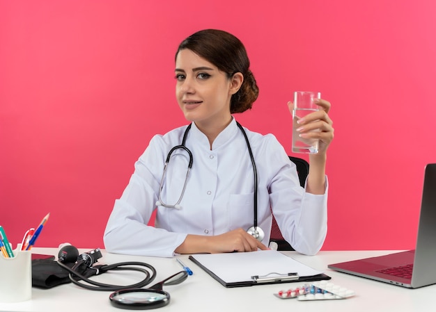 Pleased young female doctor wearing medical robe with stethoscope sitting at desk work on computer with medical tools holding glass of water with copy space