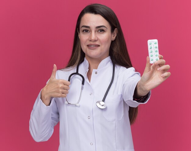 Pleased young female doctor wearing medical robe with stethoscope thumbs up
