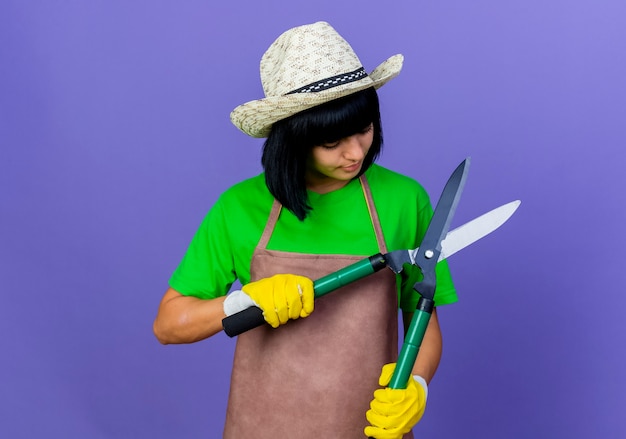 Pleased young female gardener in uniform wearing gardening hat and gloves holds and looks at clippers 