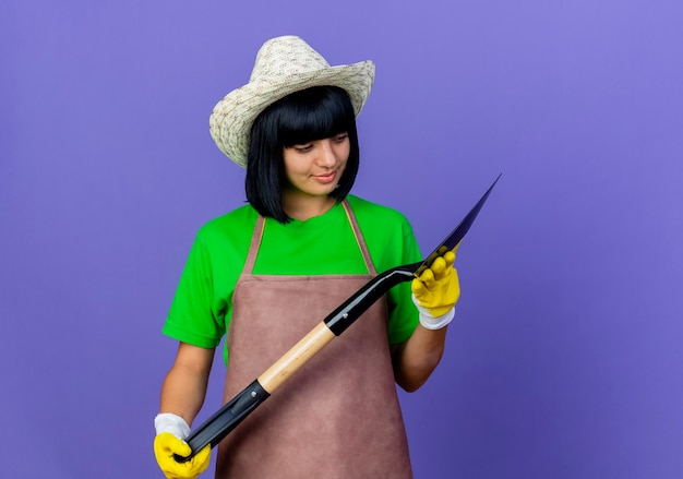 Pleased young female gardener in uniform wearing gardening hat and gloves holds and looks at spade 