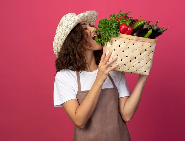 ピンクで隔離の肩に野菜のバスケットを保持し、見てガーデニング帽子を身に着けている制服を着た若い女性の庭師を喜ばせる