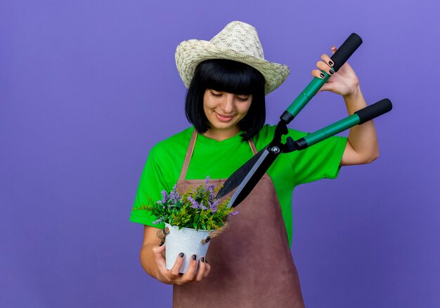 Pleased young female gardener in uniform wearing gardening hat holds clippers over flowers in flowerpot 