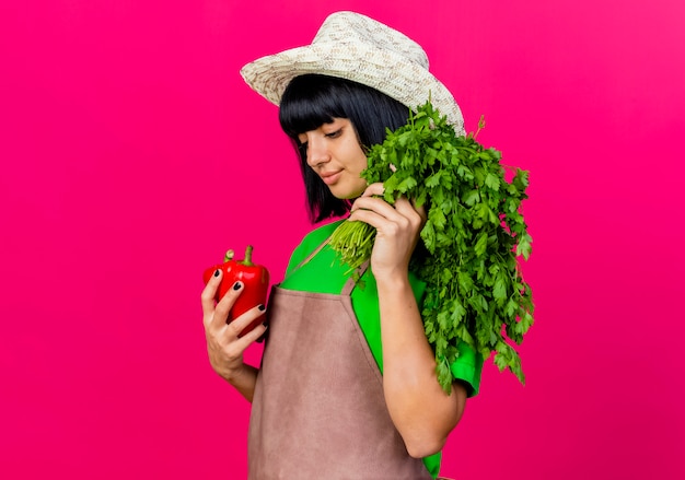 Free photo pleased young female gardener in uniform wearing gardening hat holds coriander and looks at red peppers