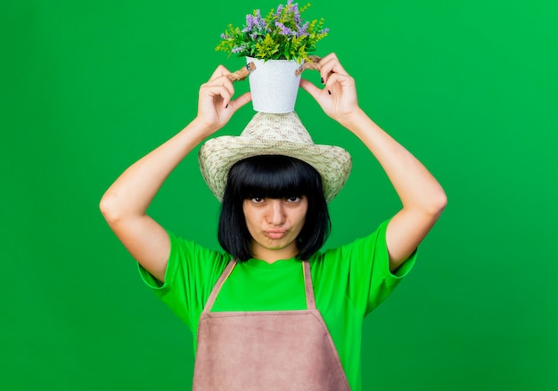 Free photo pleased young female gardener in uniform wearing gardening hat holds flowerpot over head