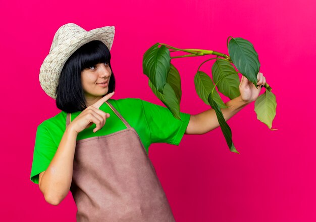 Довольная молодая женщина-садовник в униформе в садовой шляпе держит и указывает на растение