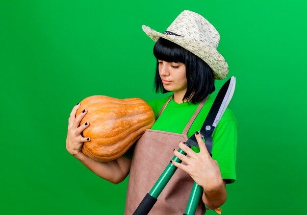 Pleased young female gardener in uniform wearing gardening hat holds pumpkin