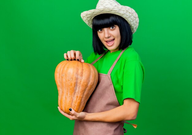 Pleased young female gardener in uniform wearing gardening hat holds pumpkin