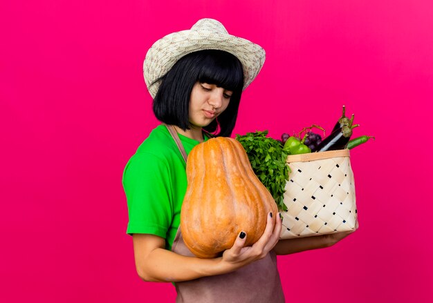 ガーデニング帽子をかぶって制服を着た若い女性の庭師は、野菜のバスケットを保持し、カボチャを見て喜んでいます