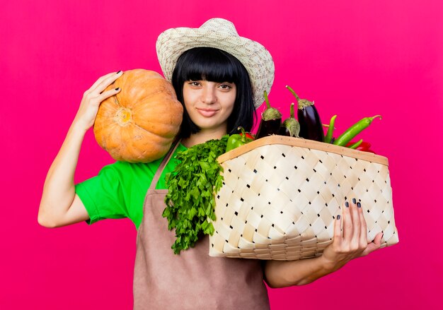 Pleased young female gardener in uniform wearing gardening hat holds vegetable basket and pumpkin 