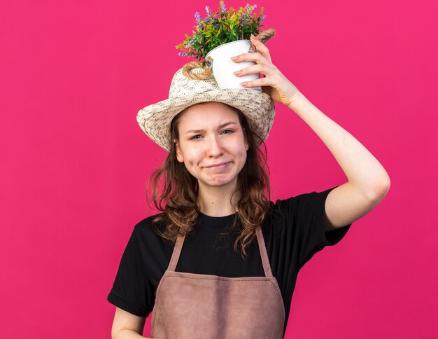 분홍색 벽에 격리된 머리에 화분에 꽃을 들고 정원용 모자를 쓴 젊은 여성 정원사를 기쁘게 생각합니다.