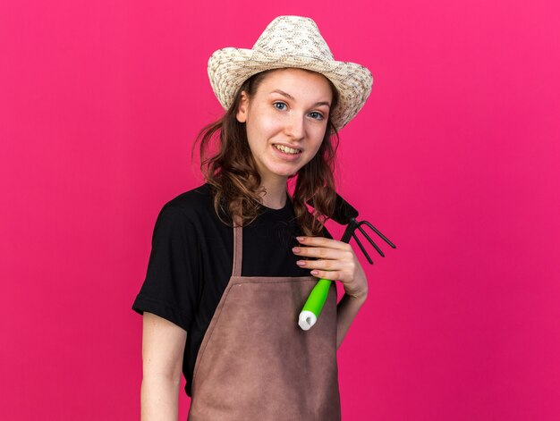 Pleased young female gardener wearing gardening hat holding hoe rake 