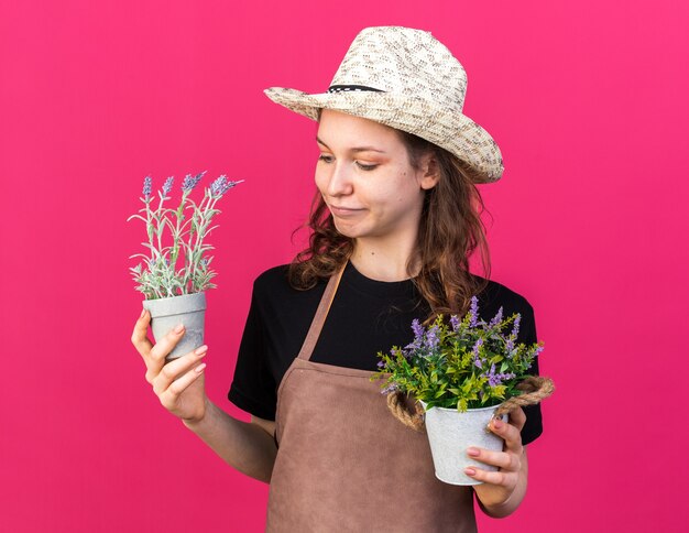 Pleased young female gardener wearing gardening hat holding and looking at flowers in flowerpots 