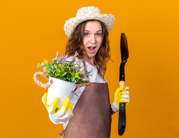 Pleased young female gardener wearing gardening hat with gloves holding flower in flowerpot with spade 