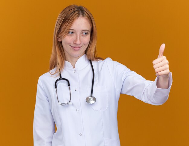 pleased young female ginger doctor wearing medical robe and stethoscope looking at side showing thumb up isolated on orange wall